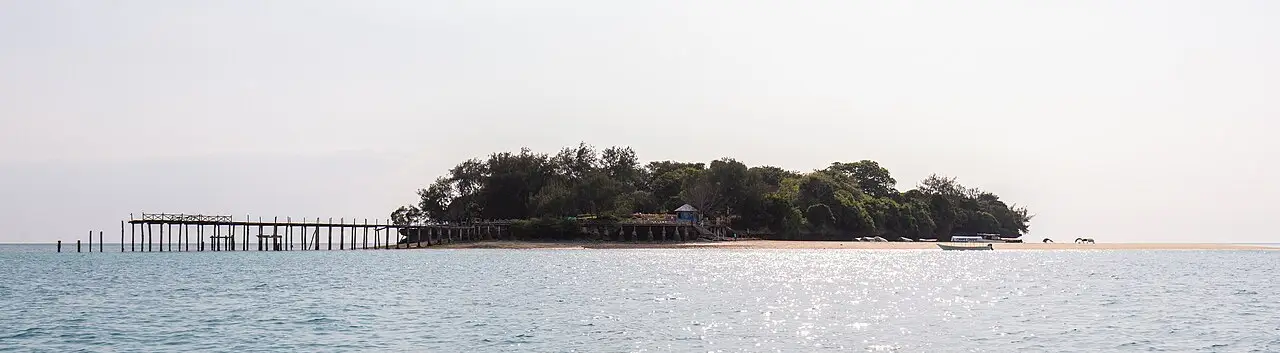 A ilha de Changuu, uma faixa baixa de árvores sobre o mar calmo, com um píer de madeira comprido à esquerda e um barco pequeno atracado à direita.
