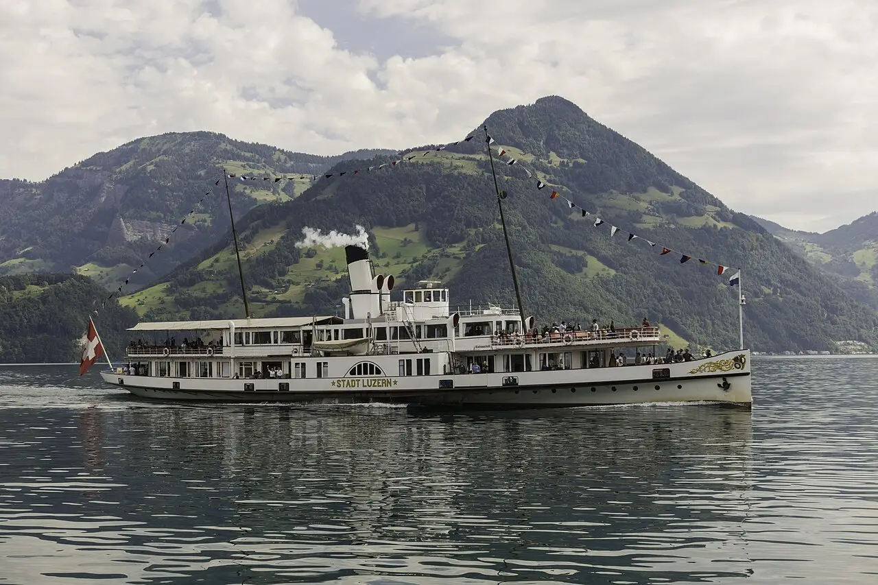 O vapor de rodas Stadt Luzern navegando no lago dos Quatro Cantões, com passageiros no convés, bandeirolas e colinas verdes ao fundo.