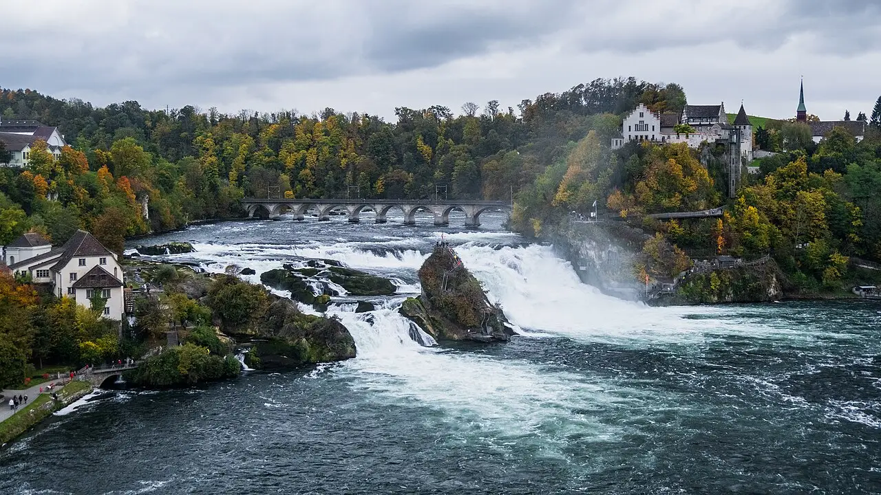 As cataratas do Reno, com a água branca caindo entre rochedos, uma ponte de arcos ao fundo, o castelo de Laufen à direita e floresta em cores de outono.