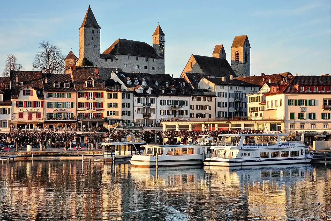 O porto de Rapperswil ao entardecer, com barcos brancos atracados, casas coloridas na orla e o castelo de torres pontudas no alto, refletidos na água.