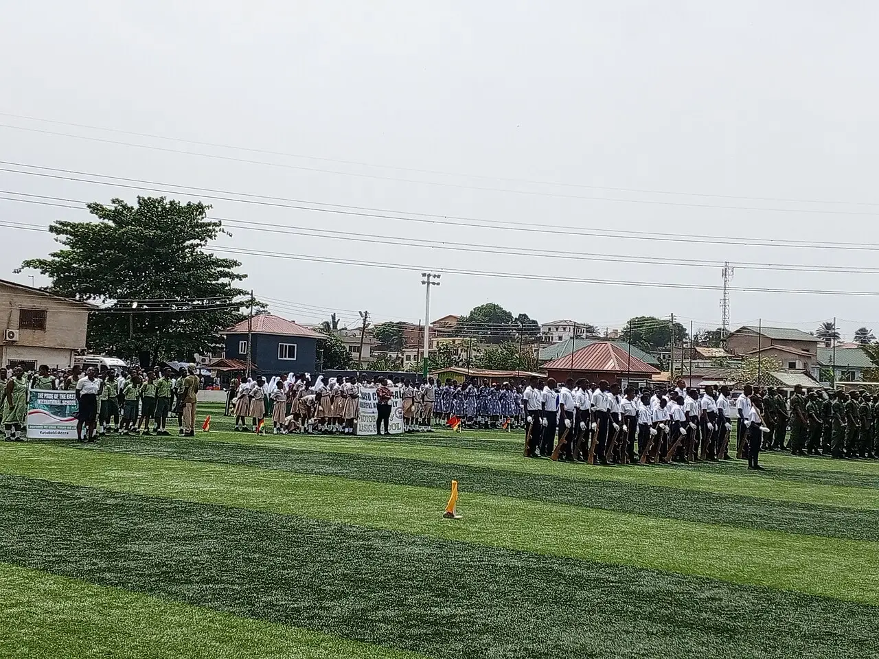 Desfile do Dia da Independência em Alajo, Acra, com grupos uniformizados marchando em um campo gramado sob céu nublado.