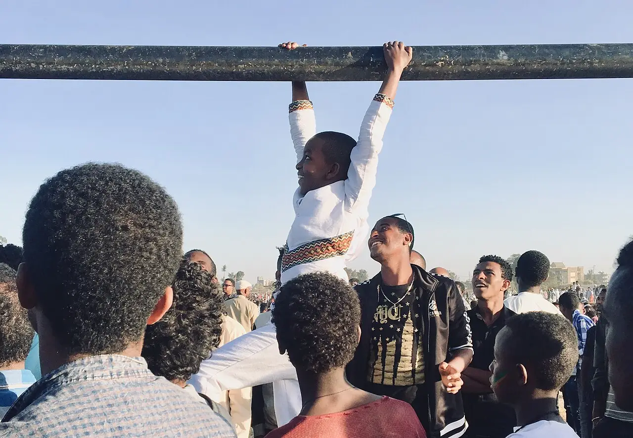 Multidão durante a celebração do Timkat em Adis Abeba, com um homem pendurado em uma viga de madeira acima das pessoas.