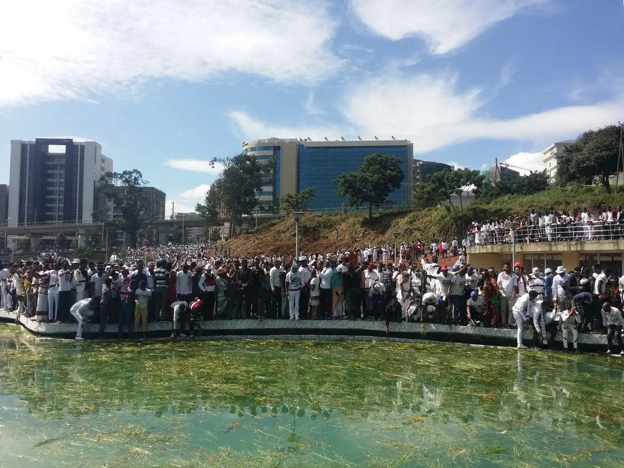 Grande multidão reunida à beira de um lago artificial durante o Irreecha, com prédios modernos ao fundo e céu azul com nuvens.