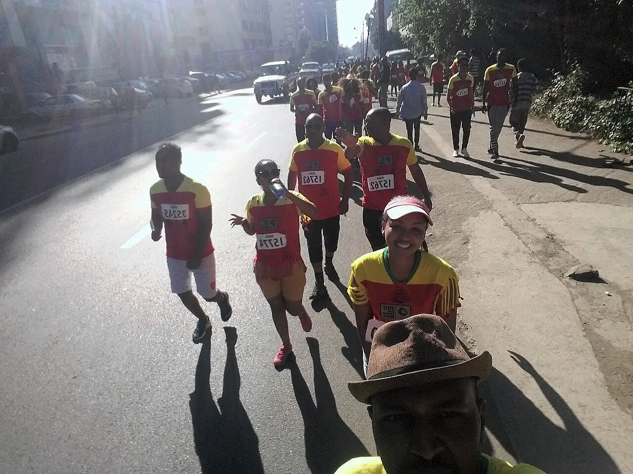 Corredores com camisetas vermelhas e números de peito descem uma rua da cidade na Great Ethiopian Run, com sombras longas no asfalto.