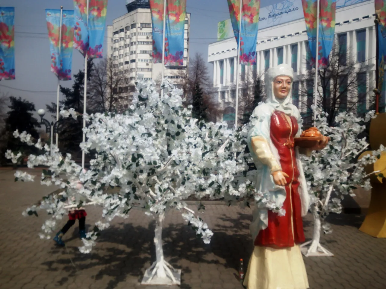 Árvores de flores brancas e uma estátua de mulher com um pão, entre bandeiras, na Praça da República de Almaty, em 23 de março de 2017.