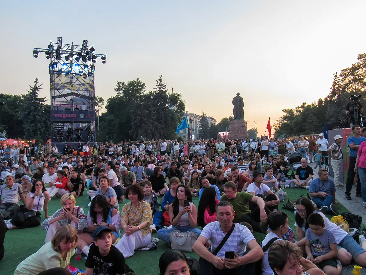 Público sentado na grama diante de um palco com torre de iluminação, em fim de tarde, no The Spirit of Tengri 2024, em Almaty.