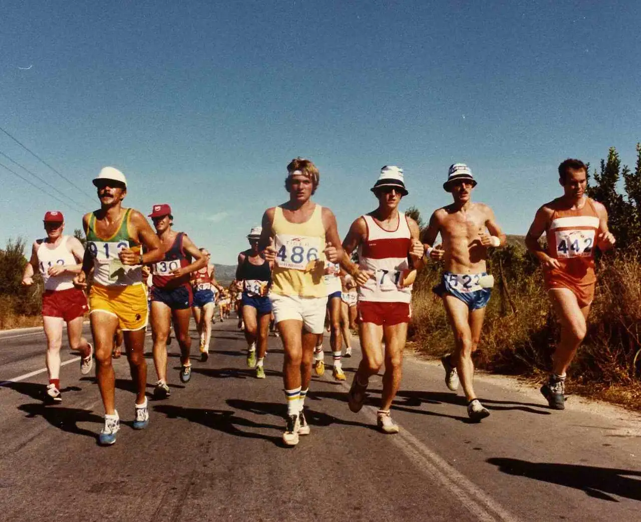 Corredores de short e camiseta numa estrada, com números no peito, na Maratona de Atenas de 1980.