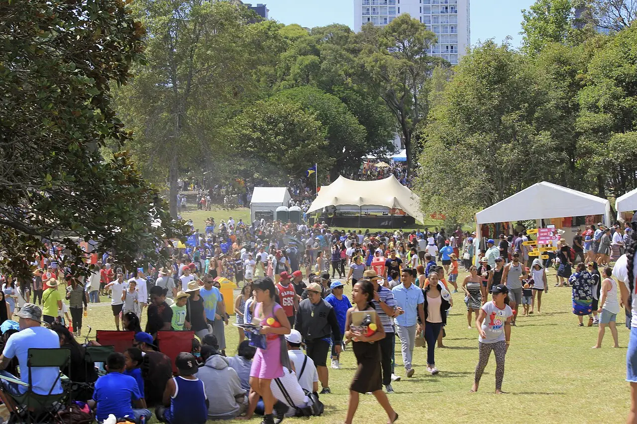 Multidão caminhando e sentada em um parque, com tendas brancas ao fundo, no Pasifika Festival de 2015, em Auckland.