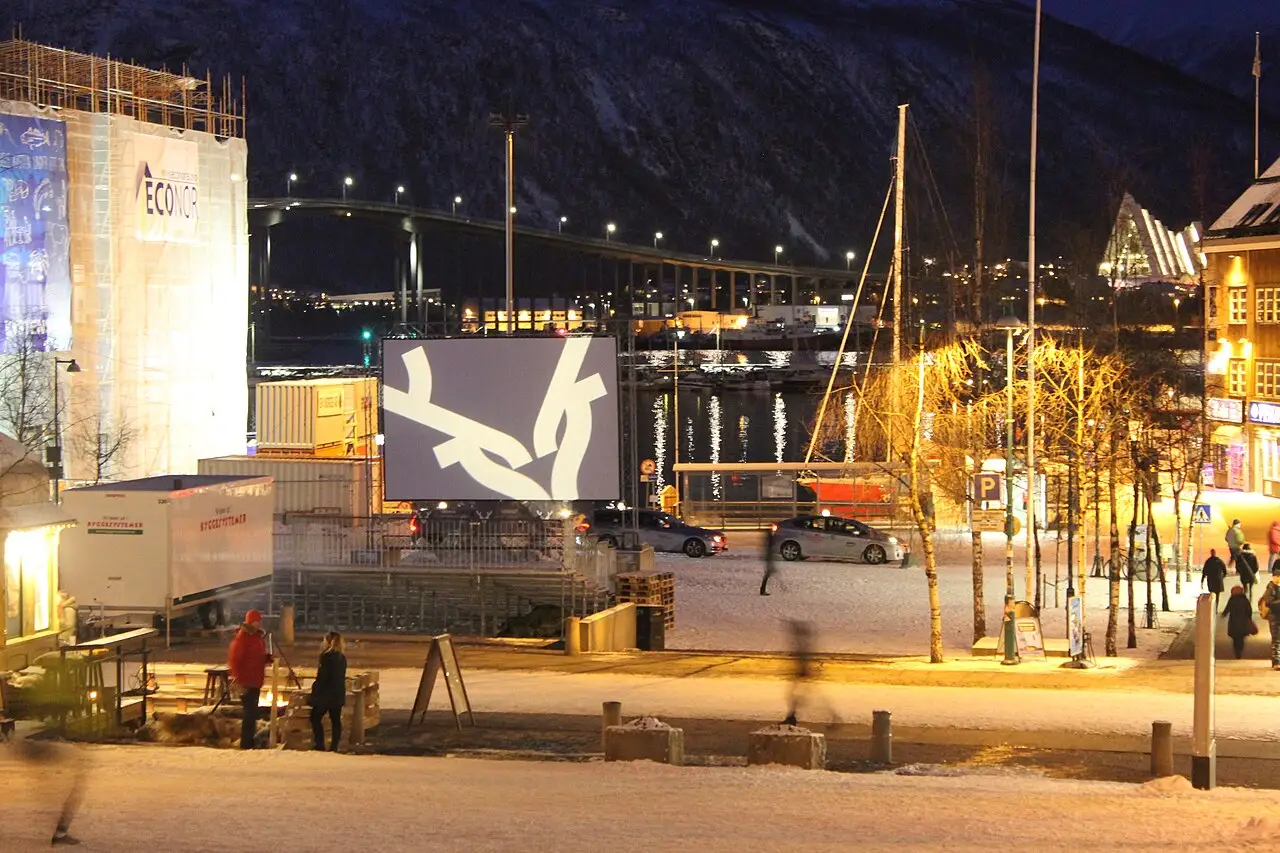 Tela de cinema ao ar livre no cais de Tromsø à noite, com a ponte e barcos iluminados ao fundo, durante o TIFF.