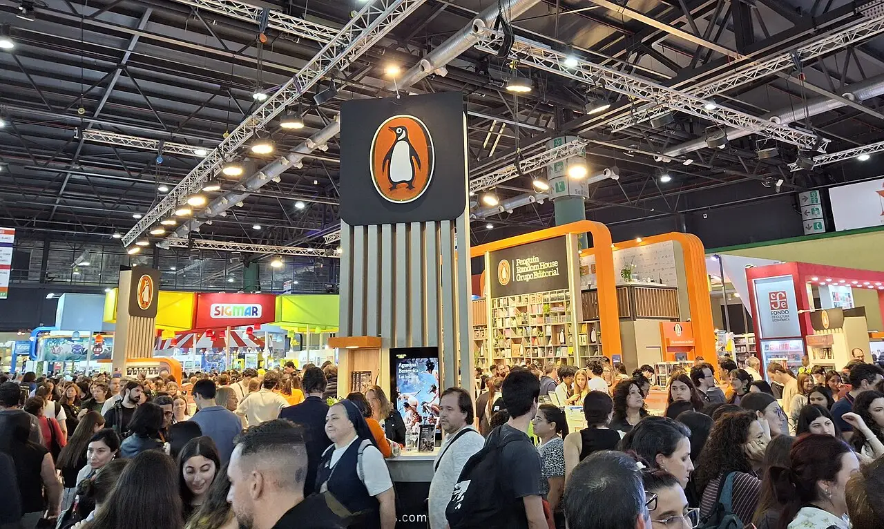 Multidão circulando entre estandes iluminados sob a estrutura metálica de um pavilhão na Feria Internacional del Libro de Buenos Aires de 2025.
