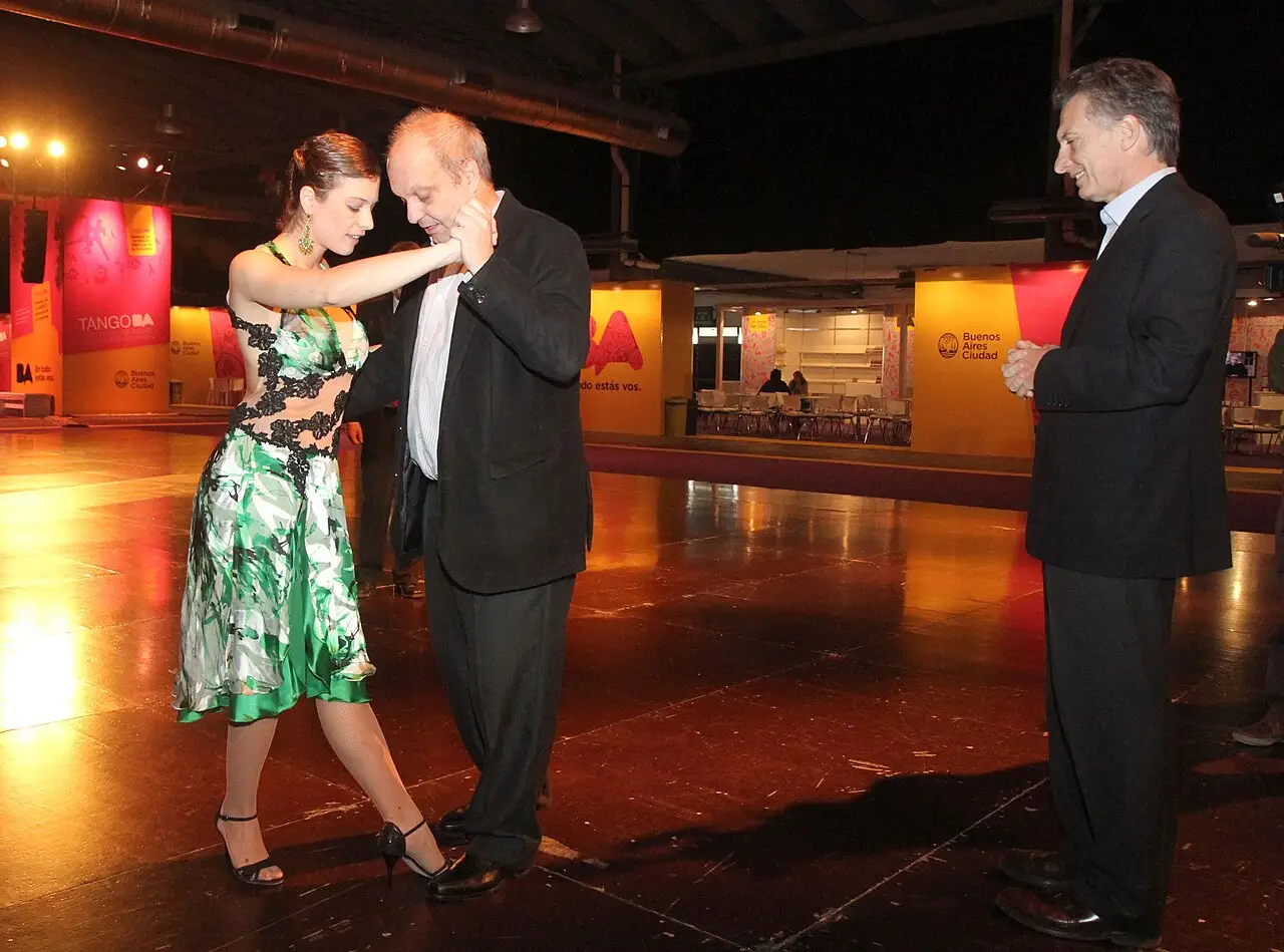 Uma mulher de vestido verde estampado dança tango com um homem em um salão de exposições, com painéis da cidade de Buenos Aires ao fundo.