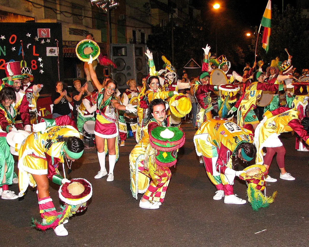 Grupo de murga com fantasias coloridas, tambores e adereços na rua à noite durante o Carnaval porteño, em Buenos Aires.