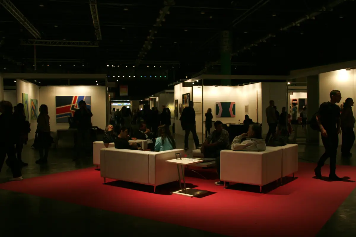 Sala de feira de arte com estandes iluminados, sofás brancos sobre um tapete vermelho e visitantes ao fundo na arteBA de 2011.