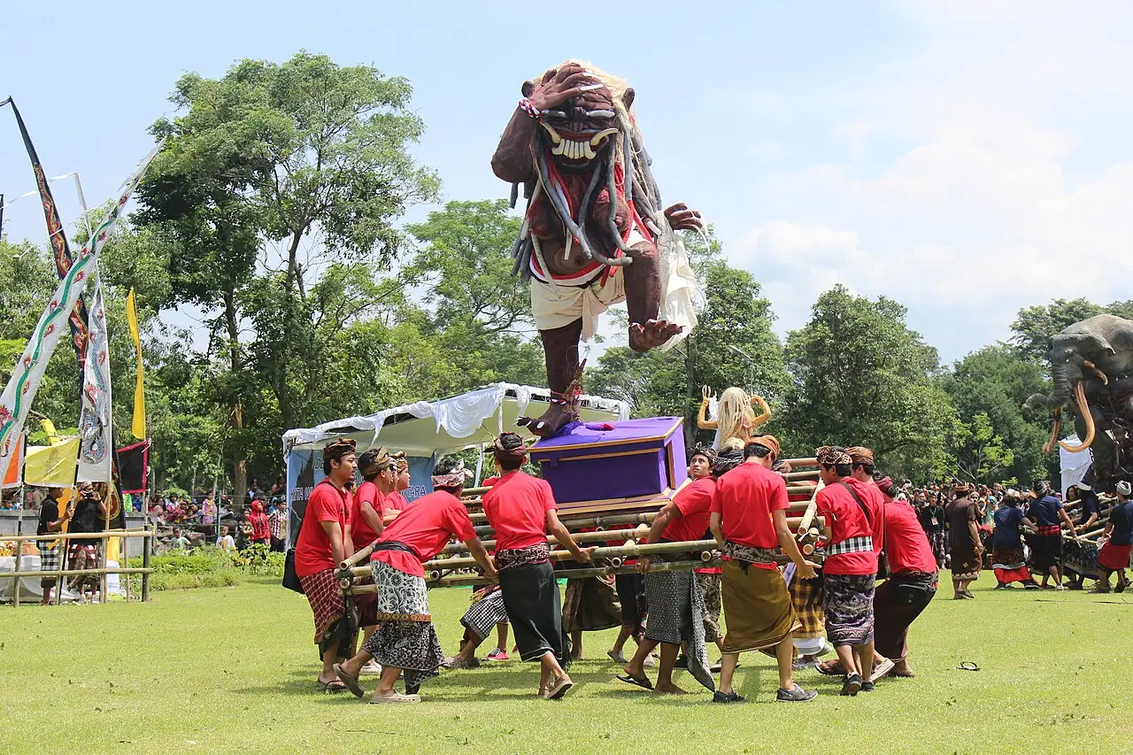 Homens de camisa vermelha carregam uma figura ogoh-ogoh sobre uma plataforma em um gramado durante as festividades do Nyepi, em Bali.