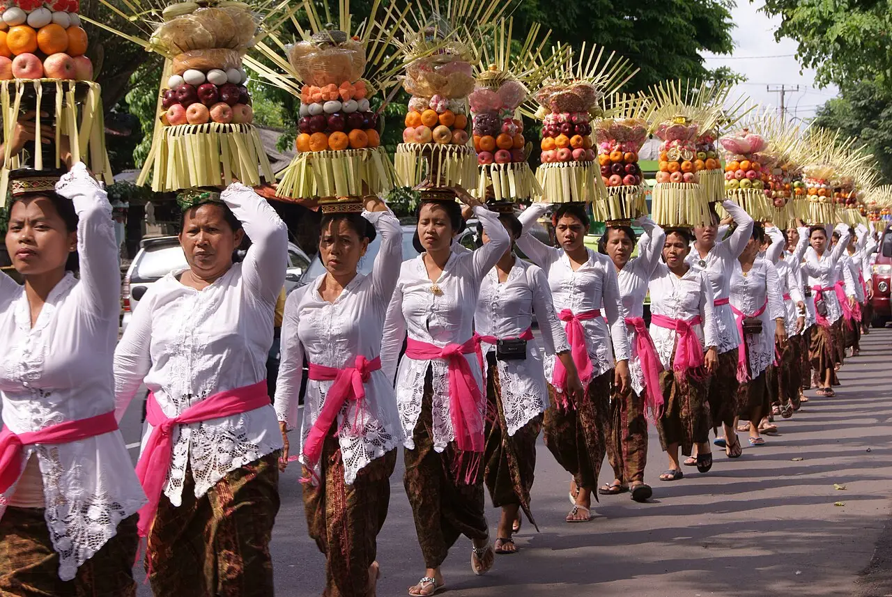 Fila de mulheres de branco com faixas rosa carregando oferendas altas na cabeça em procissão por uma estrada durante um odalan.