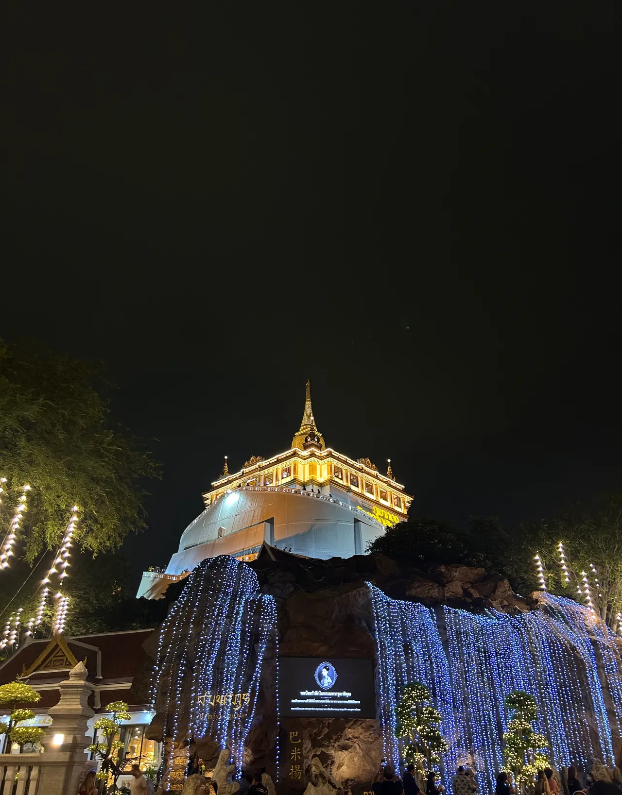 O chedi da Montanha Dourada, no Wat Saket, iluminado à noite em 30 de outubro de 2025, com cortinas de luz azul na rocha e gente pequena ao pé.
