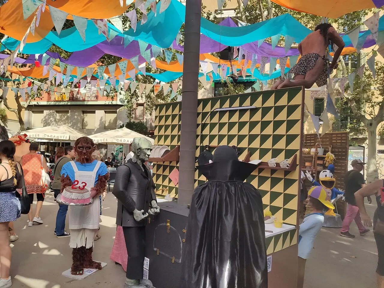 Rua decorada com bandeirolas coloridas e cenários em papel, com pessoas passeando, na Festa Major de Gràcia de 2023, em Barcelona.