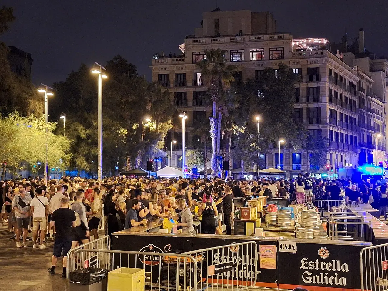 Multidão de noite na praça Universitat, em Barcelona, com mesas de comida e edifícios iluminados, na revetlla de Sant Joan de 2024.
