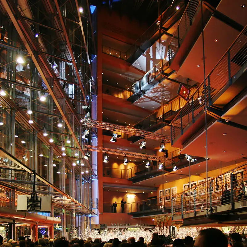 Interior do Berlinale Palast com escadas, passarelas de metal, paredes de vidro e luzes quentes, com pessoas ao fundo.