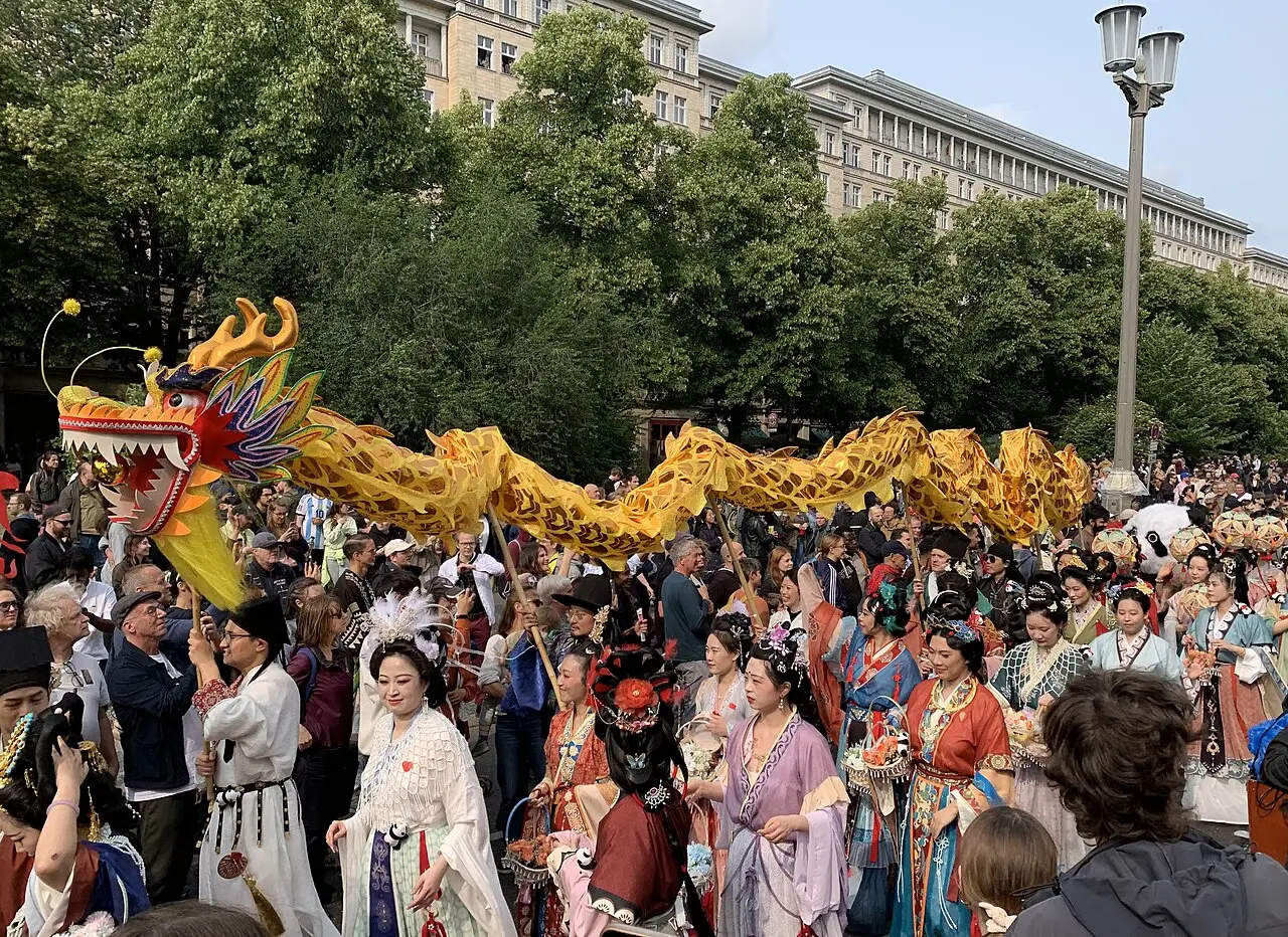 Dragão dourado de tecido conduzido por uma multidão em fantasias em uma avenida arborizada no Karneval der Kulturen de 2025, em Berlim.