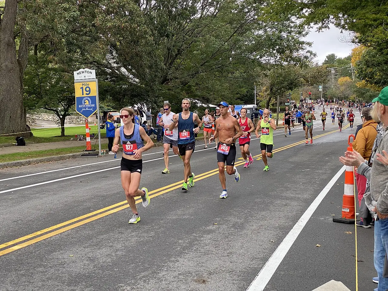 Corredores pasan por la milla 19 del Maratón de Boston de 2021 en una calle arbolada con señales de tráfico.