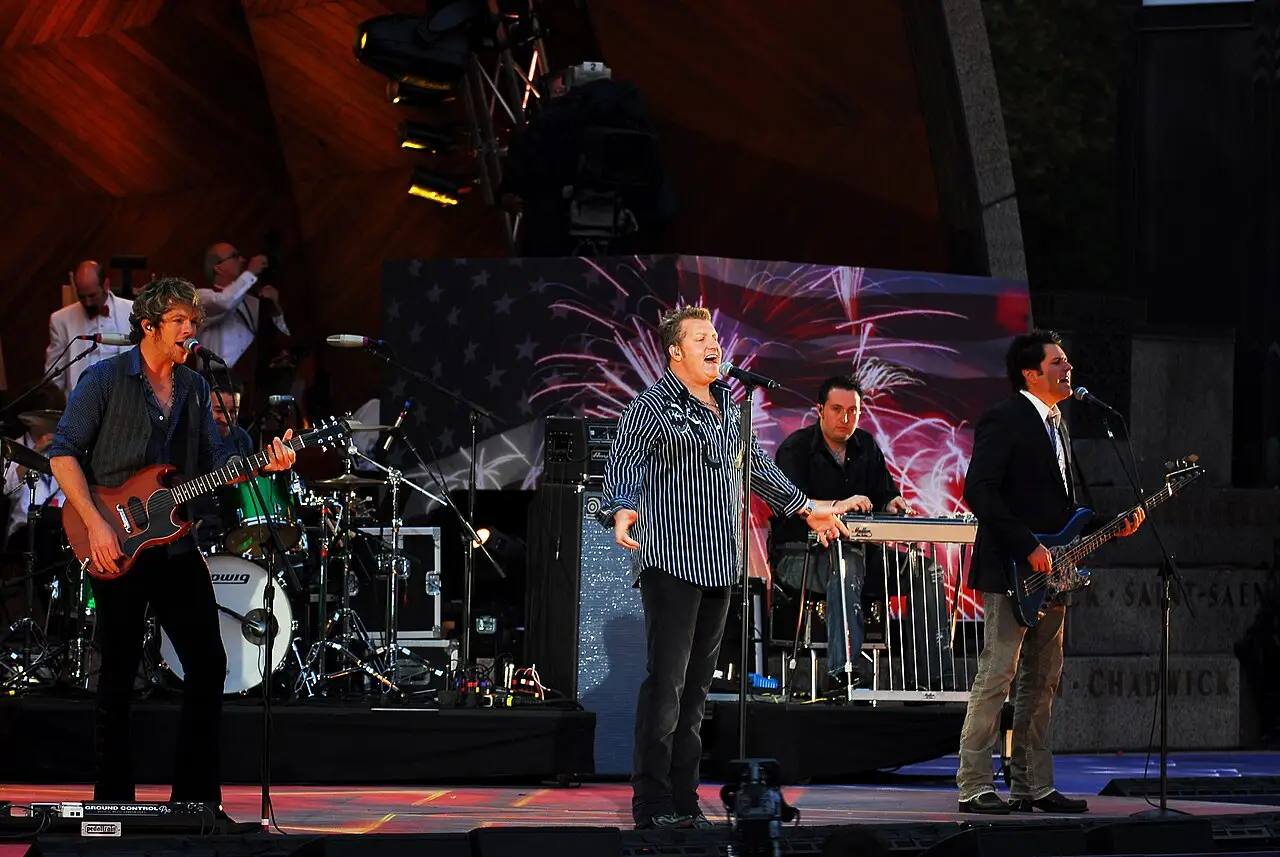 Una banda de rock en un escenario con batería y guitarras frente a una pantalla con la bandera estadounidense en el Boston Pops Fireworks Spectacular, en la Esplanade.