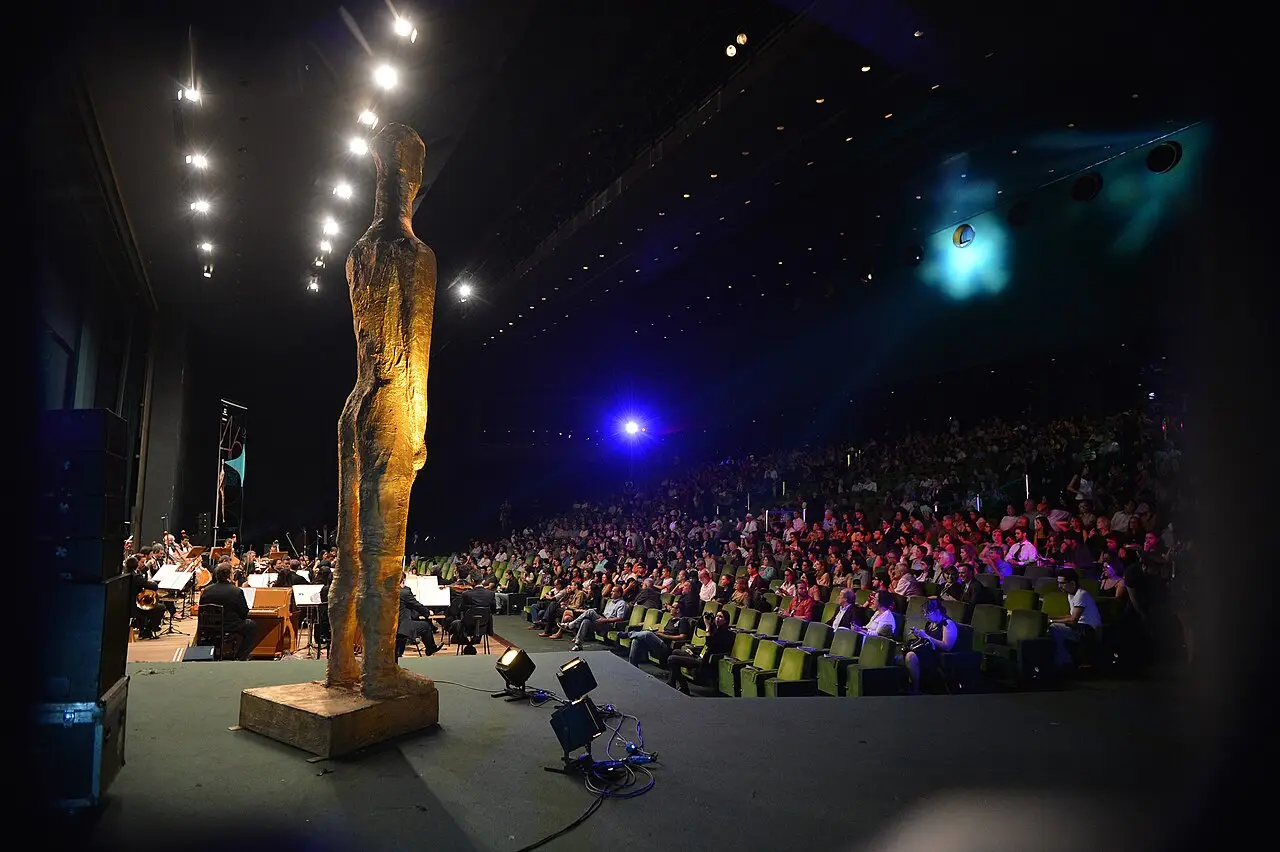 Palco com uma grande escultura dourada, músicos e uma plateia iluminada em azul e violeta, na abertura do festival de 2013.