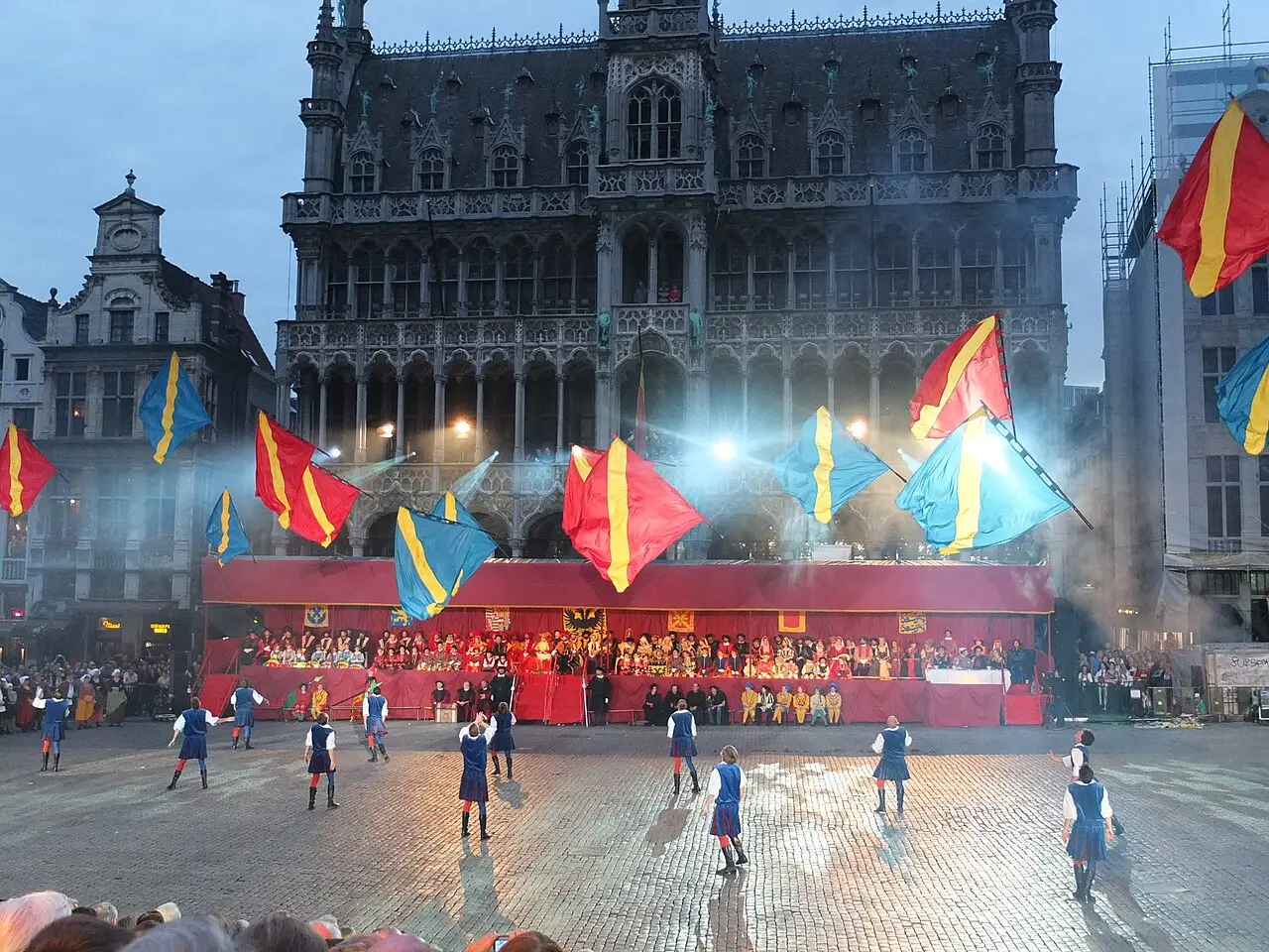 Espetáculo do Ommegang na Grand-Place de Bruxelas ao anoitecer, com plateia, bandeiras e a fachada da prefeitura ao fundo.