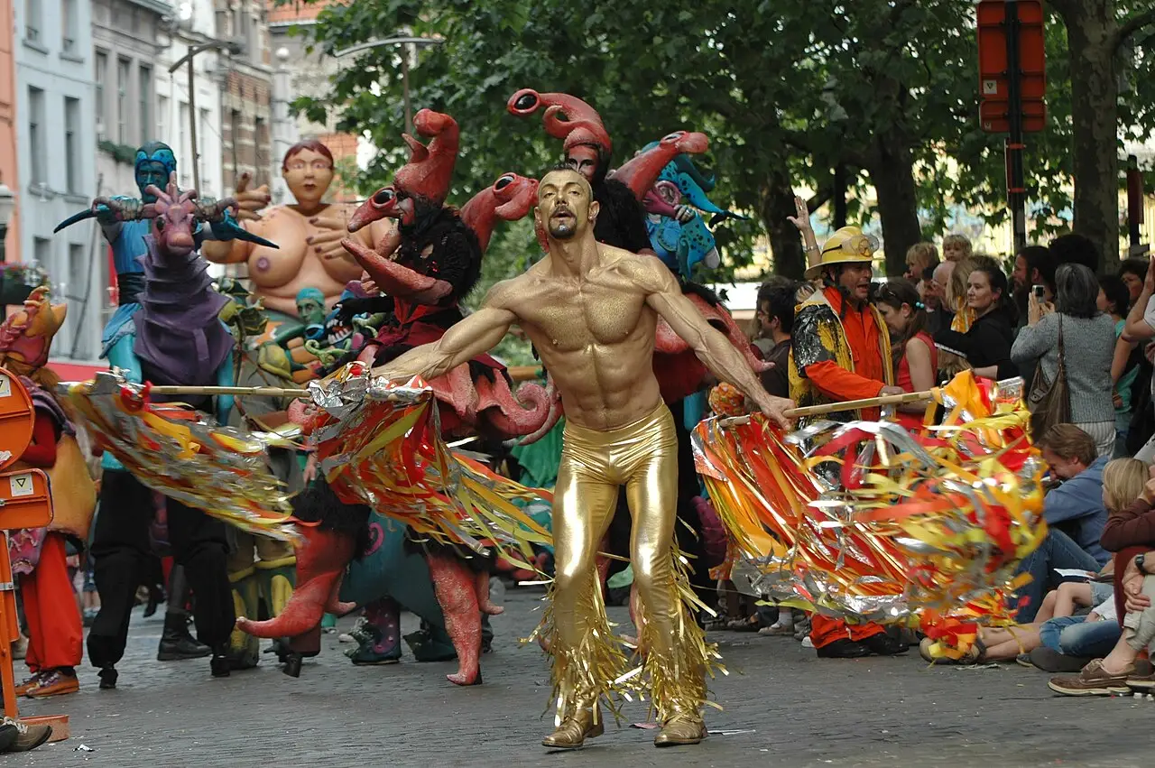 Participantes com fantasias coloridas e uma figura dourada dançando em uma rua arborizada de Bruxelas durante a Zinneke Parade.