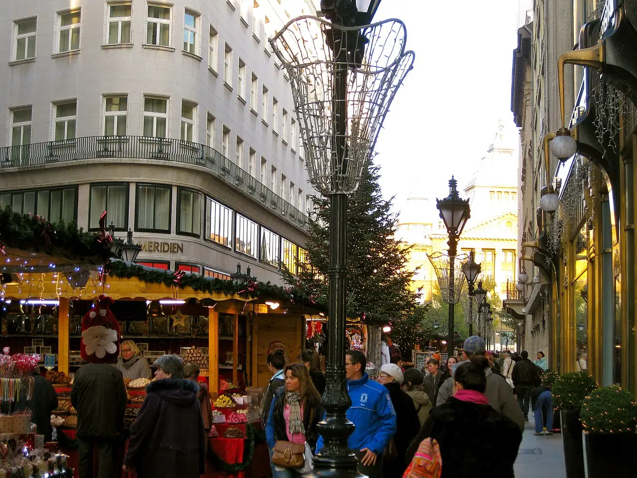 Barracas de madeira e visitantes no mercado de Natal da rua Deák Ferenc, em Budapeste, em 2012, com edifícios altos ao redor.