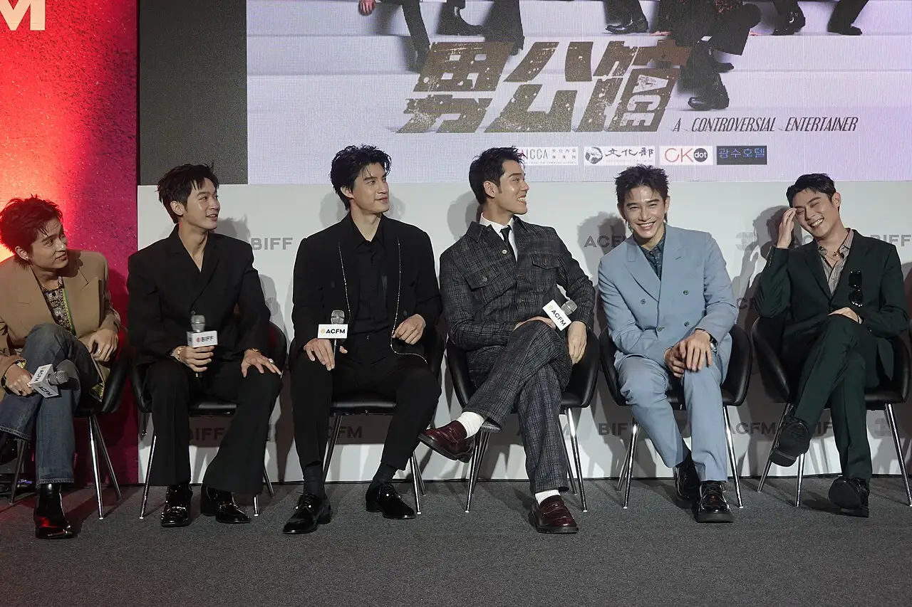 Elenco do filme A Controversial Entertainer sentado em fila diante de um painel do filme no Festival Internacional de Cinema de Busan de 2025.