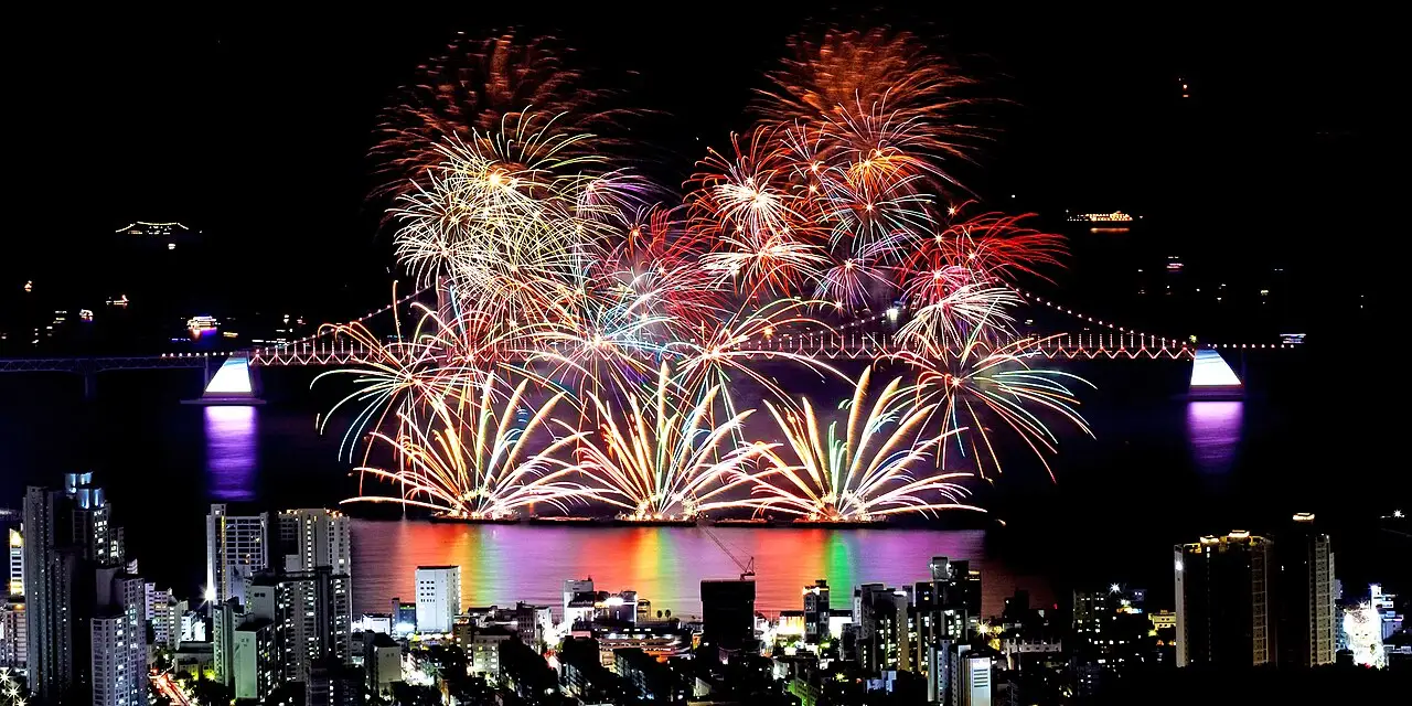 Fogos de artifício em vermelho e branco explodem sobre a ponte e os prédios da cidade, à noite, no Festival de Fogos de Busan.