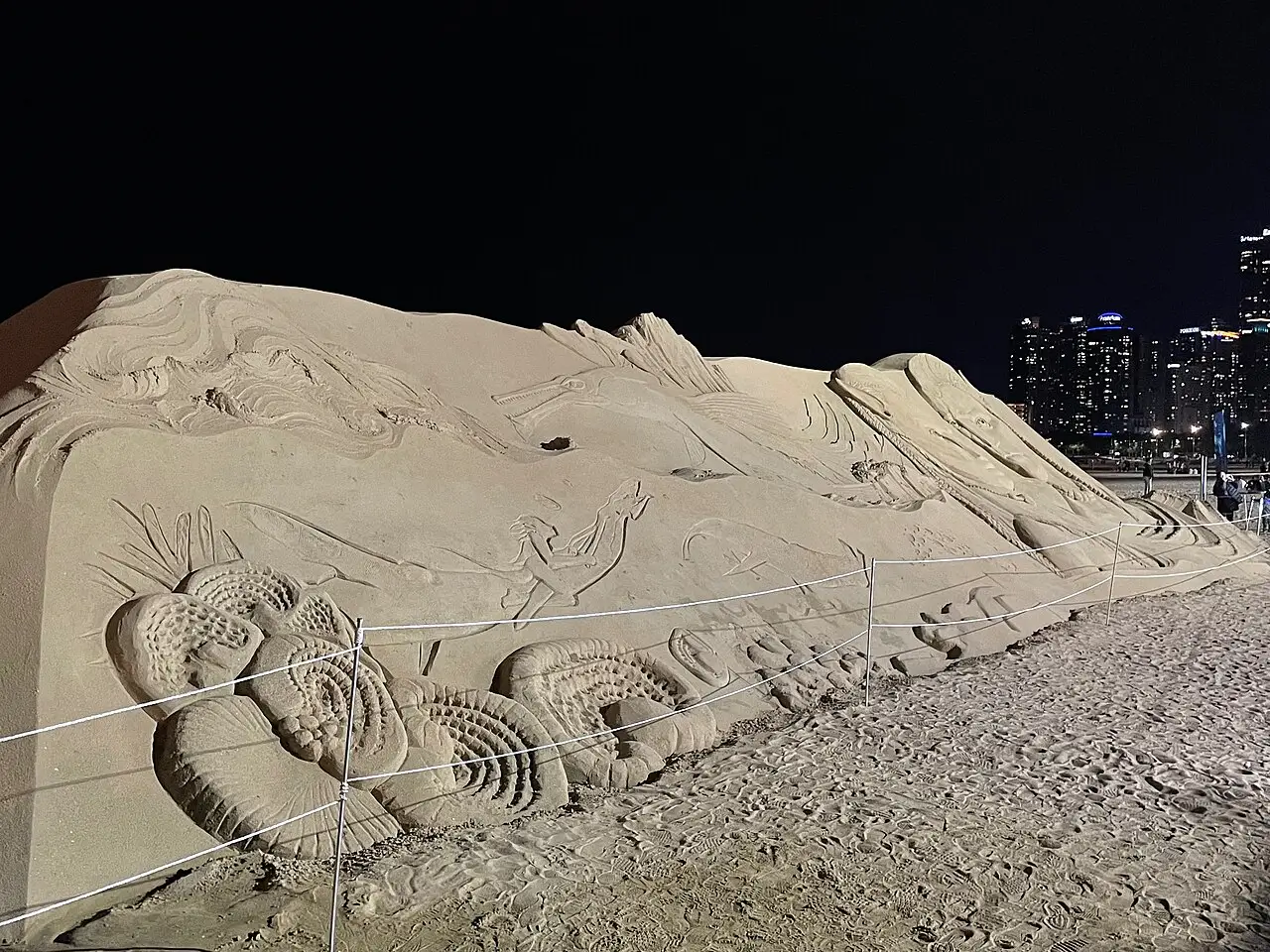 Escultura de areia com relevos de figuras na praia de Haeundae, à noite, com prédios iluminados da cidade ao fundo.