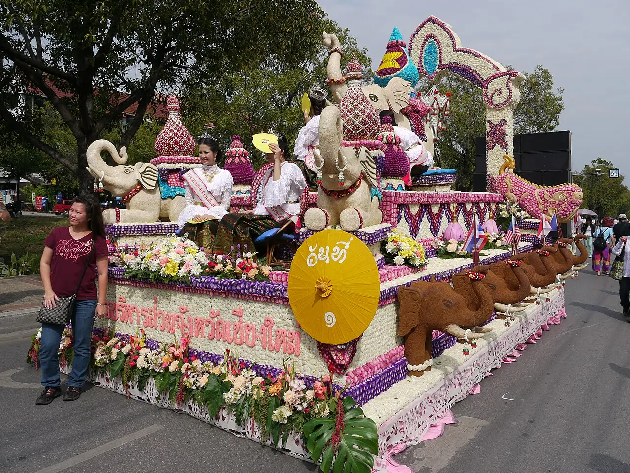Carro alegórico coberto de flores, com esculturas e pessoas em cima, desfilando por uma rua de Chiang Mai durante o Festival das Flores.
