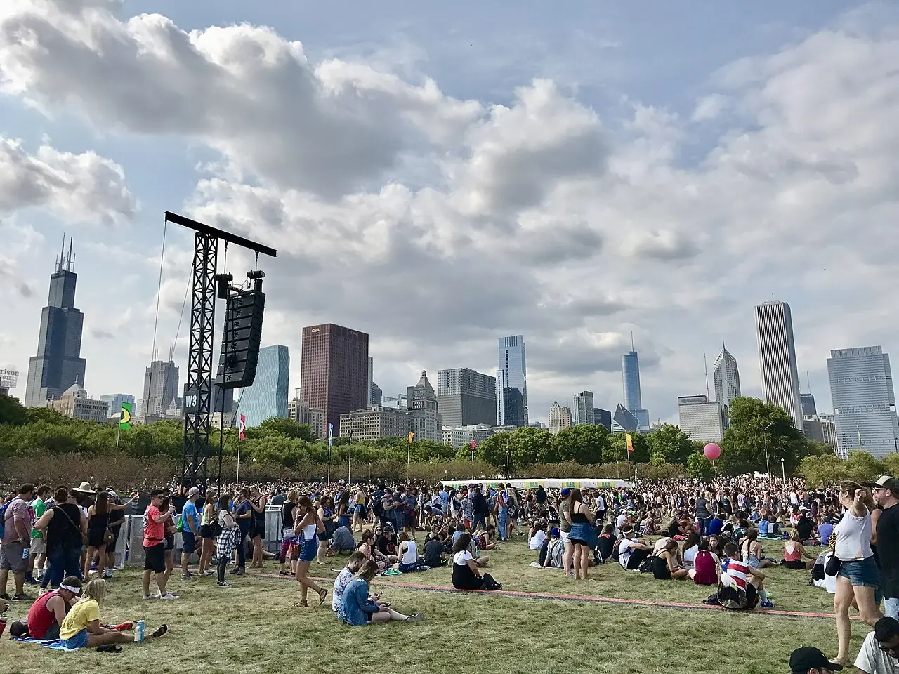 Un public assis sur l’herbe d’un parc, avec une tour de scène et les gratte-ciel de Chicago derrière, au Lollapalooza.