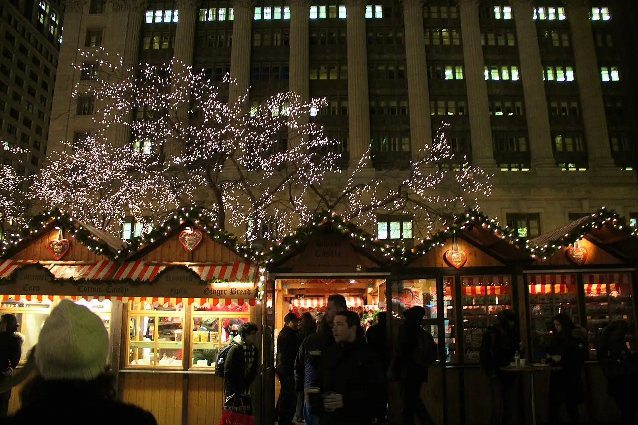 Des étals en bois éclairés du Christkindlmarket la nuit, avec un arbre couvert de lumières devant un grand immeuble à Chicago.