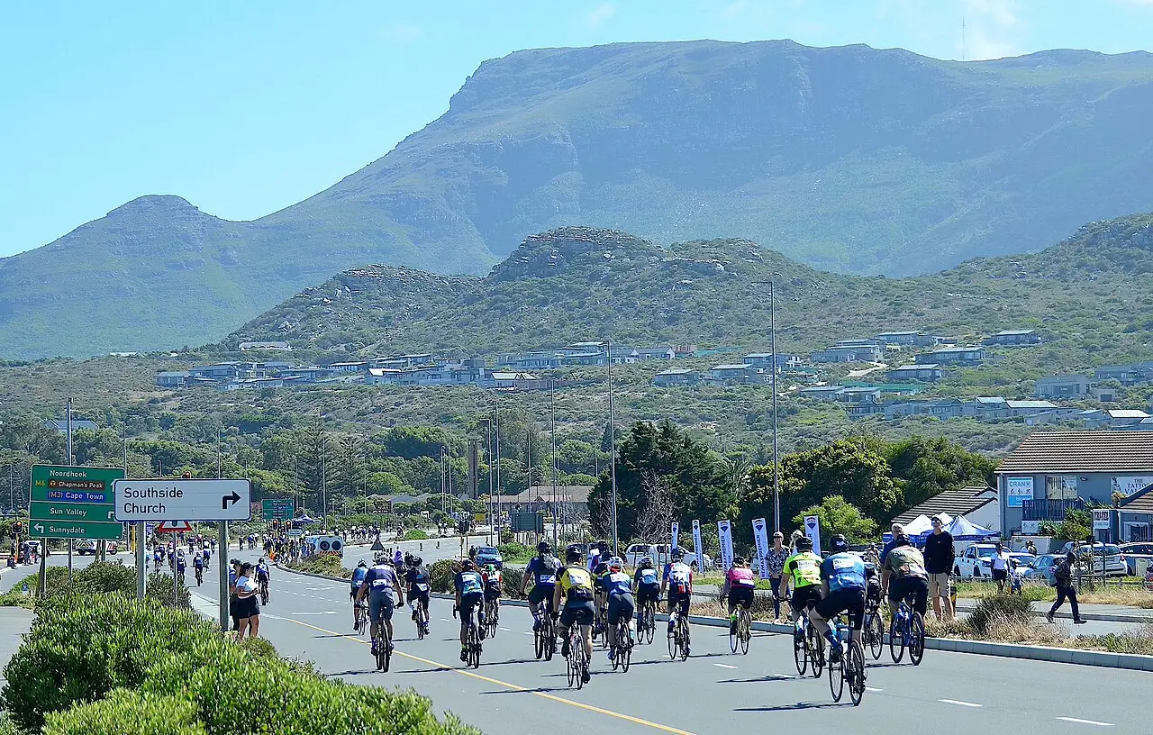 Ciclistas em grupo pedalam em uma estrada, com montanhas ao fundo sob céu azul, no Cape Town Cycle Tour de 2024.