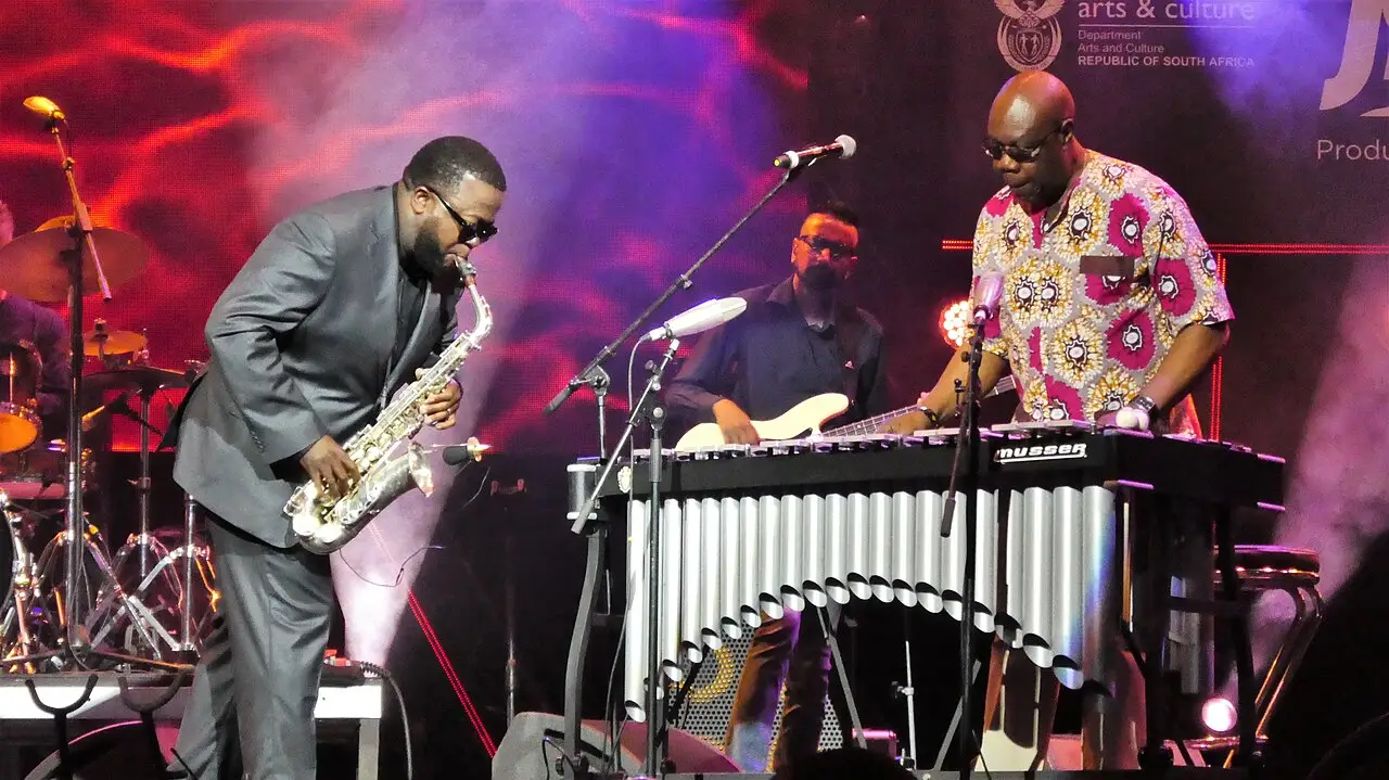 Dois músicos no palco, um de camisa estampada tocando saxofone e outro ao vibrafone, sob luzes vermelhas em um festival de jazz na Cidade do Cabo.