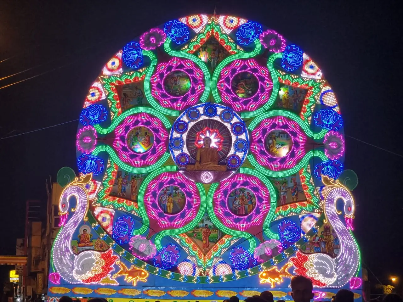 Un grand cadre lumineux circulaire, le thorana de Vesak à Grandpass, avec des motifs concentriques violets, verts et bleus allumés la nuit.