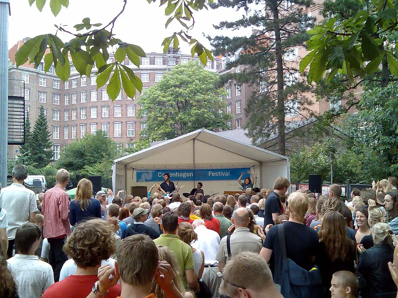 Multidão em um pátio arborizado diante de uma tenda branca do Copenhagen Jazz Festival, em Copenhague, em dia claro.
