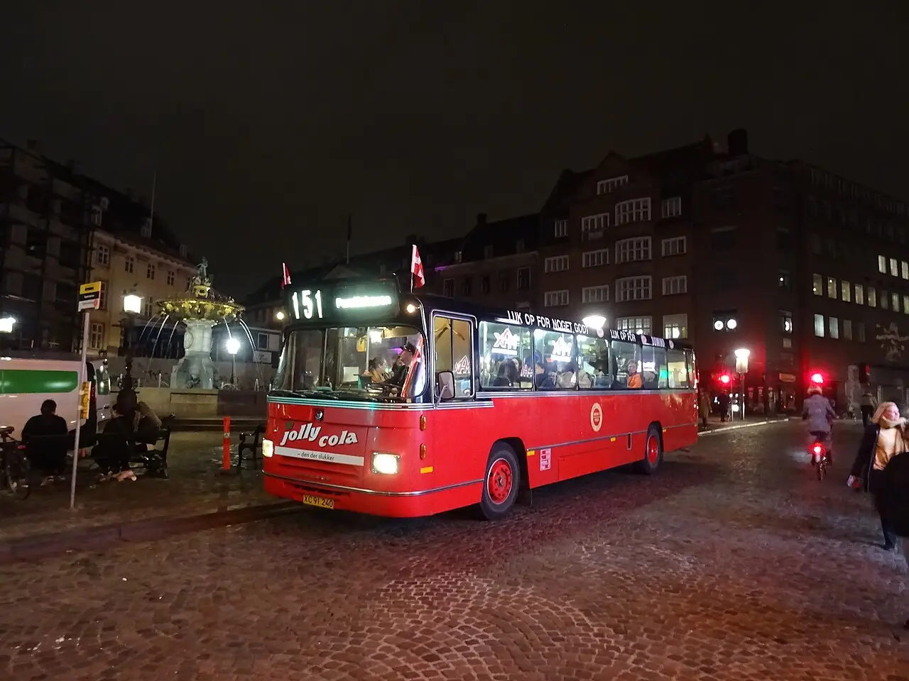 Ônibus vermelho antigo parado à noite ao lado da fonte do Gammeltorv, com bandeiras da Dinamarca, na Kulturnatten de 2022 em Copenhague.