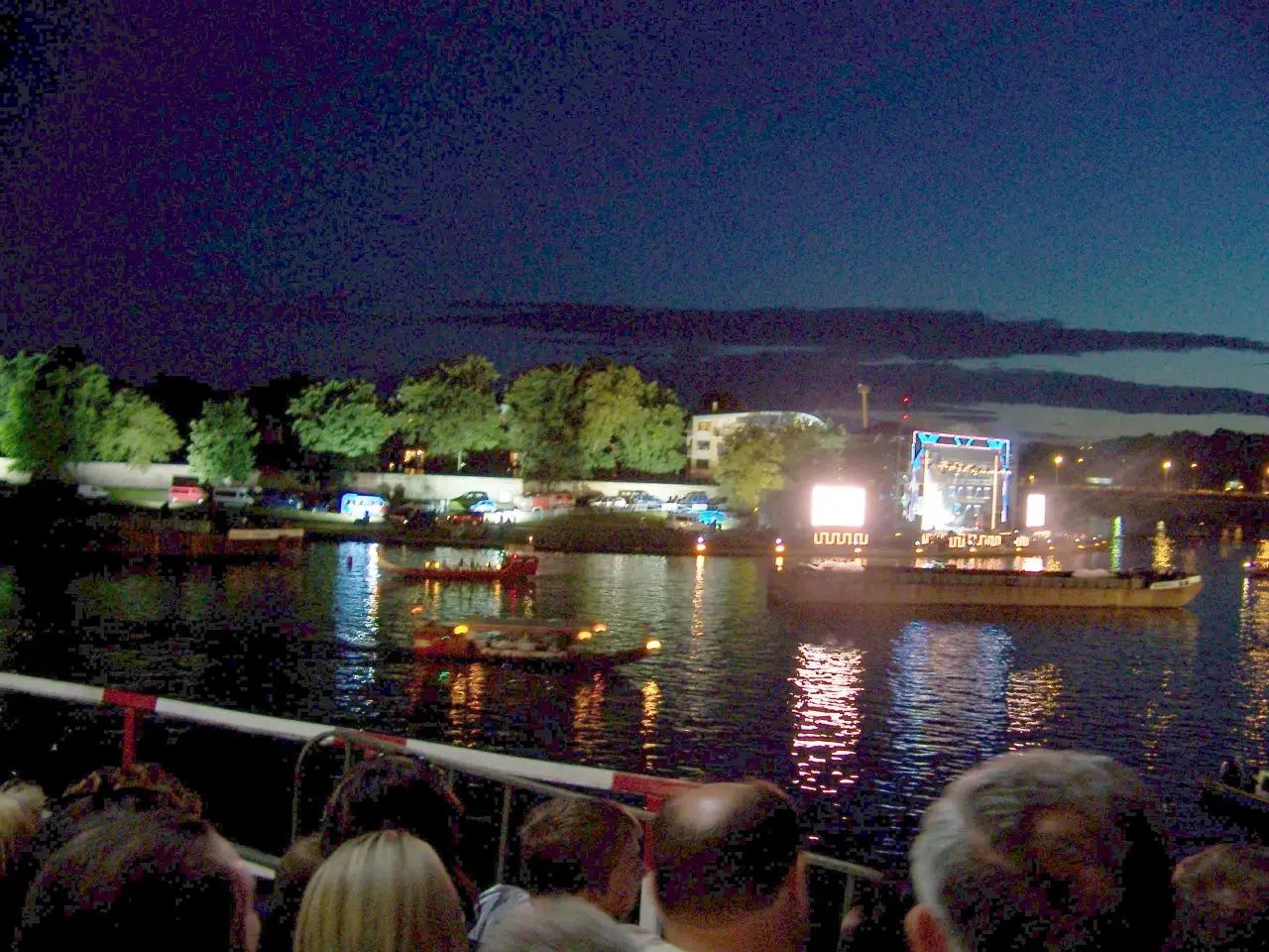 Palco iluminado sobre a água e barcos no rio à noite, visto da margem cheia de gente, no Wianki de 2007, em Cracóvia.