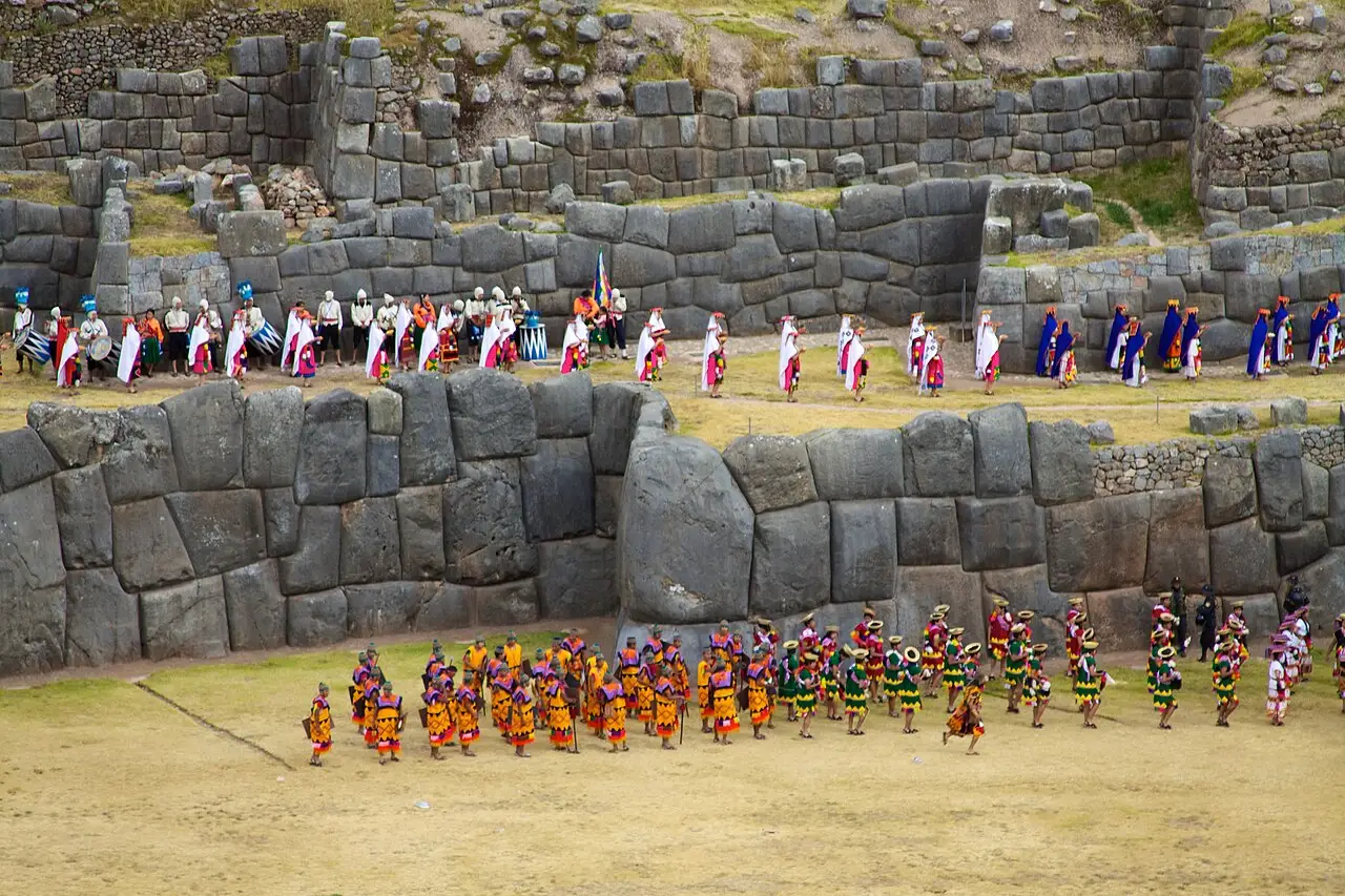 Participantes de trajes vermelhos e laranja em fileiras diante dos muros de pedra de Sacsayhuamán durante a cerimônia do Inti Raymi.