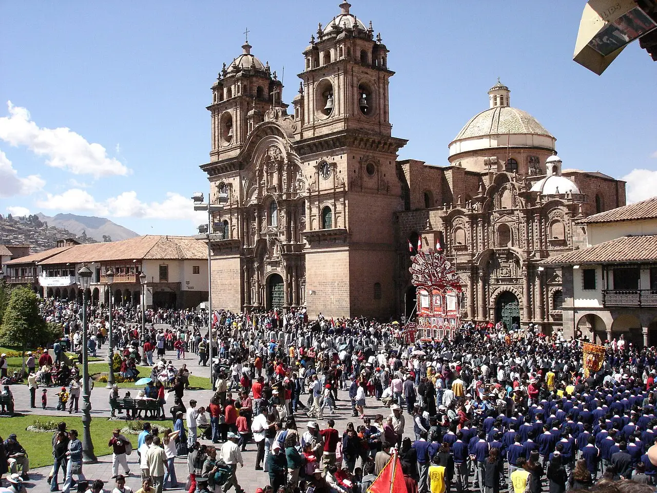 Praça cheia de gente diante de uma igreja barroca de duas torres em Cusco durante o Corpus Christi, sob céu azul.