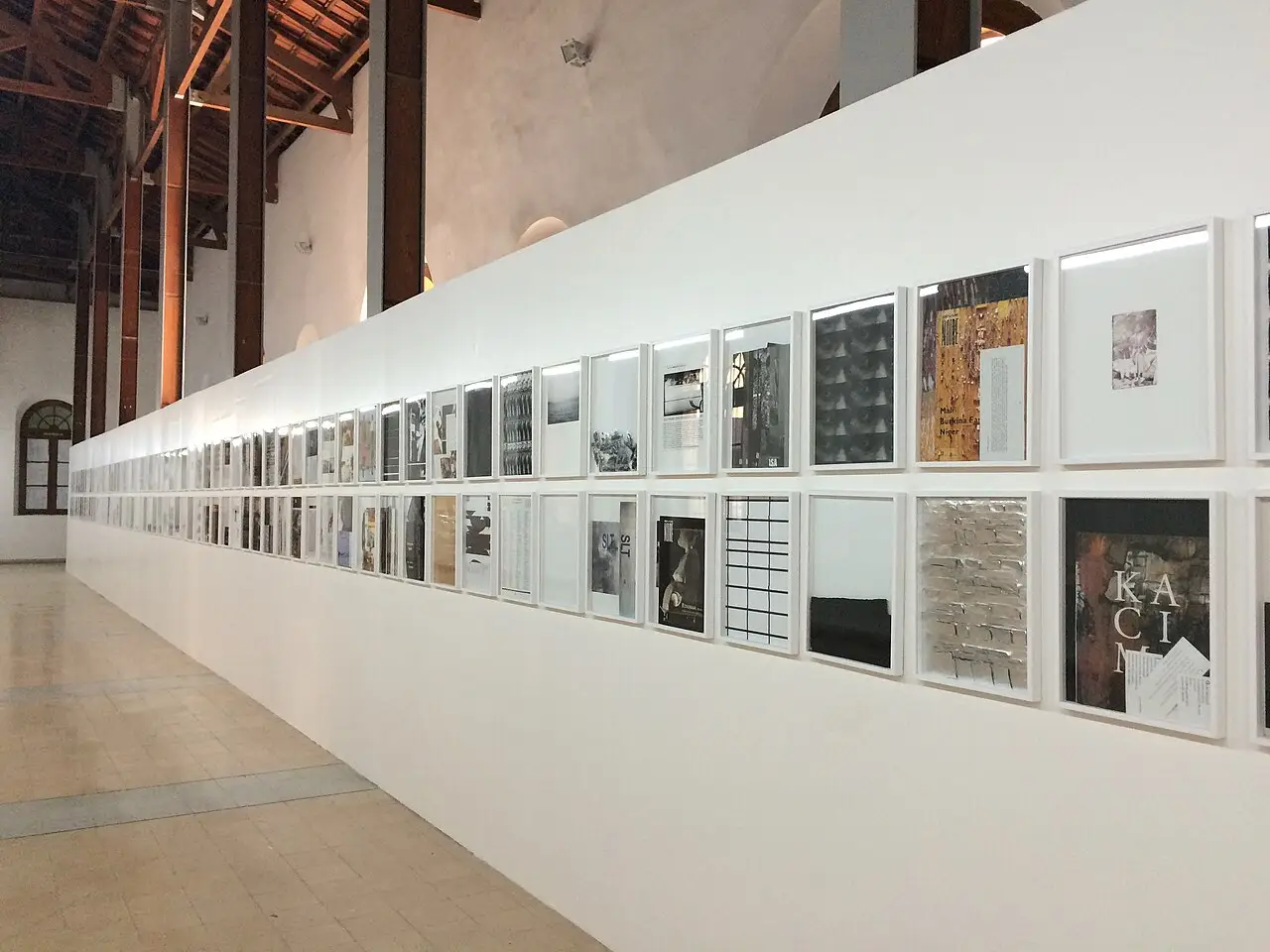Corredor de uma galeria com uma longa parede branca cheia de imagens emolduradas na Bienal Dak’Art de 2016, em Dacar.