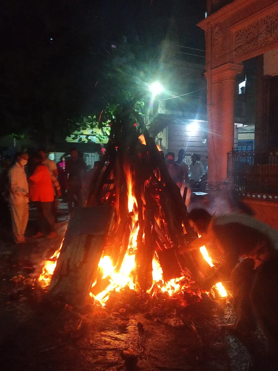 Fogueira de troncos acesa à noite em Delhi, com pessoas ao fundo, na véspera do Holi de 2021.