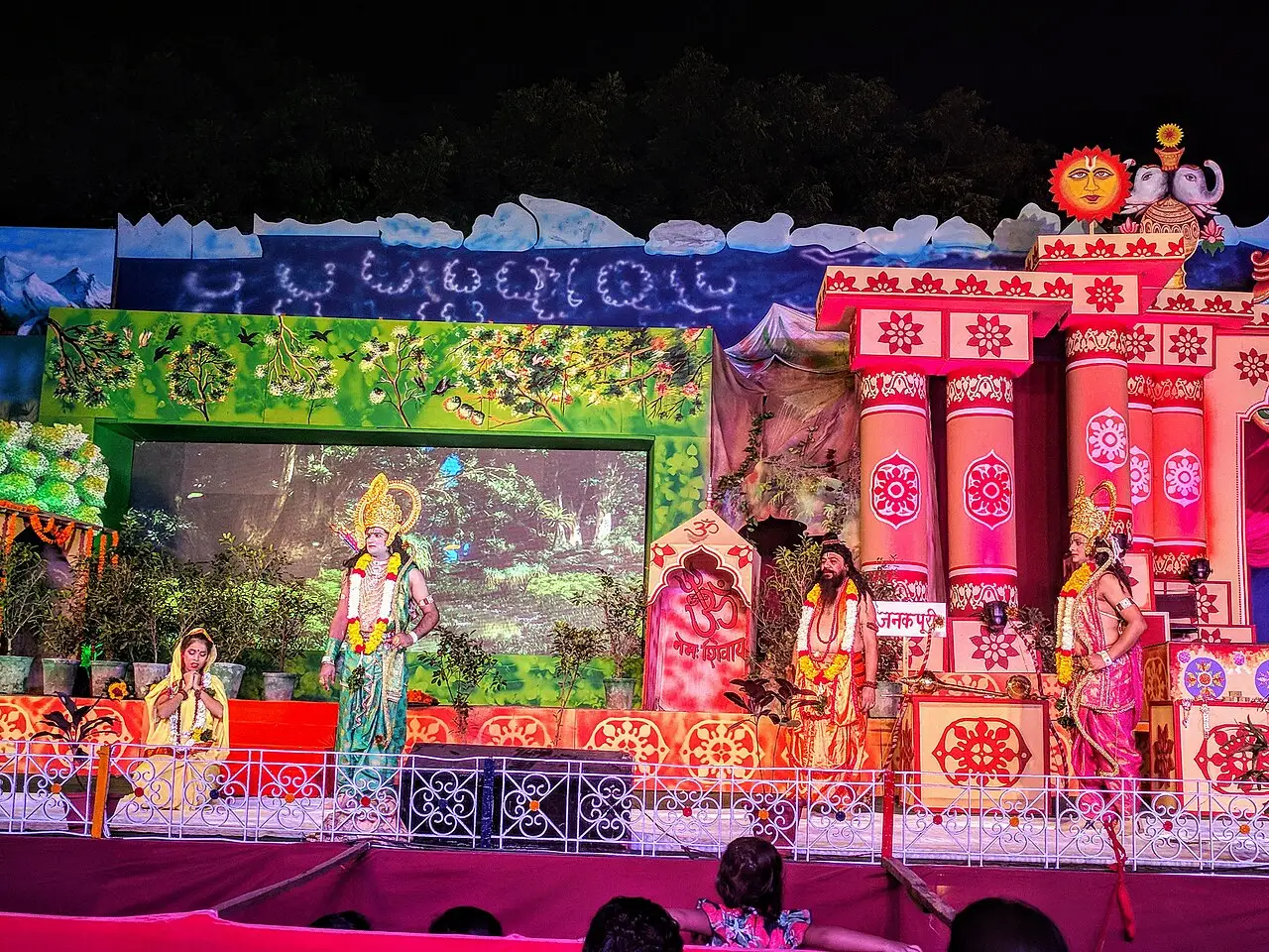 Palco iluminado e decorado em cores vivas, com atores em trajes tradicionais encenando o Ramlila diante do Forte Vermelho, à noite.