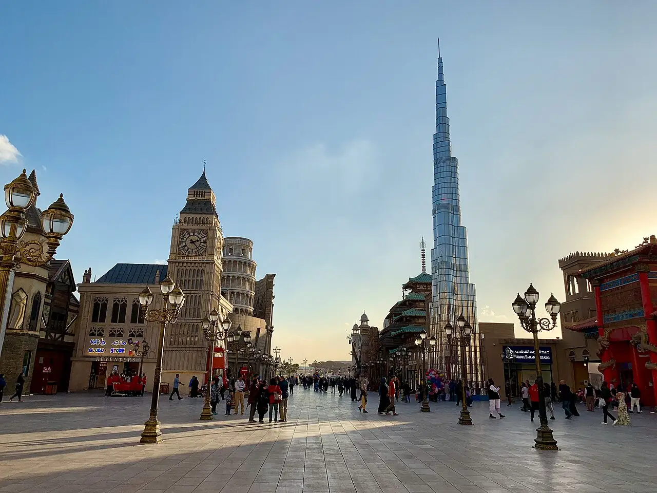 Praça com pavilhões decorados e postes de luz no Global Village, em Dubai, com visitantes caminhando e uma torre alta ao fundo, em fevereiro de 2019.