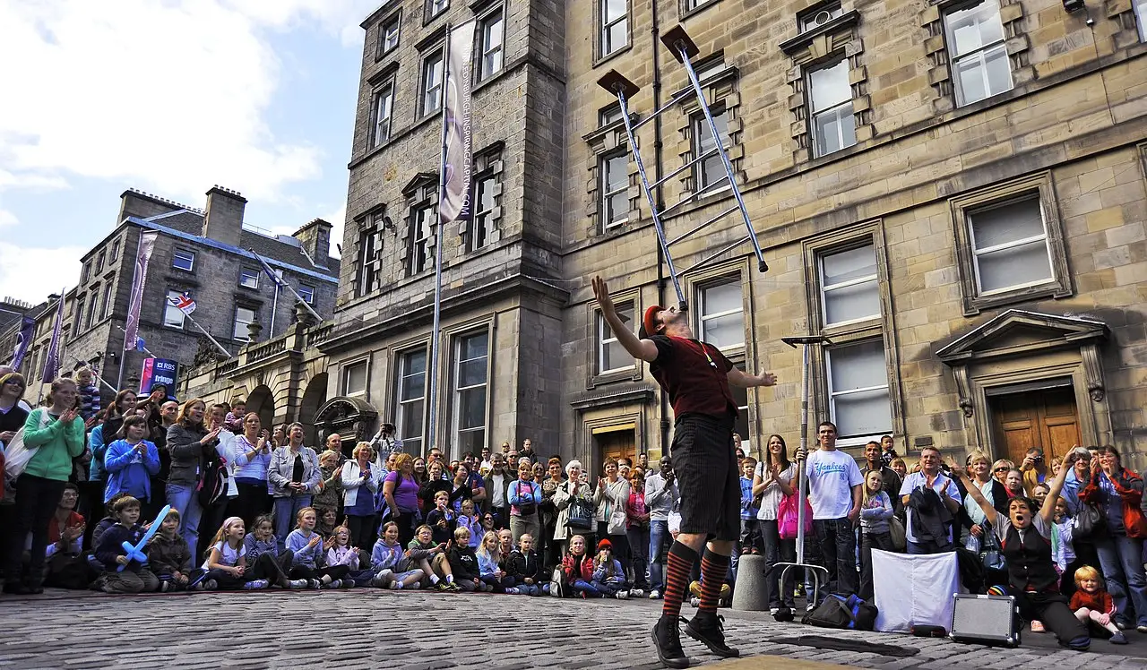 Artista de rua em pé diante de uma plateia sentada no calçamento de uma rua histórica de Edimburgo durante o Fringe.