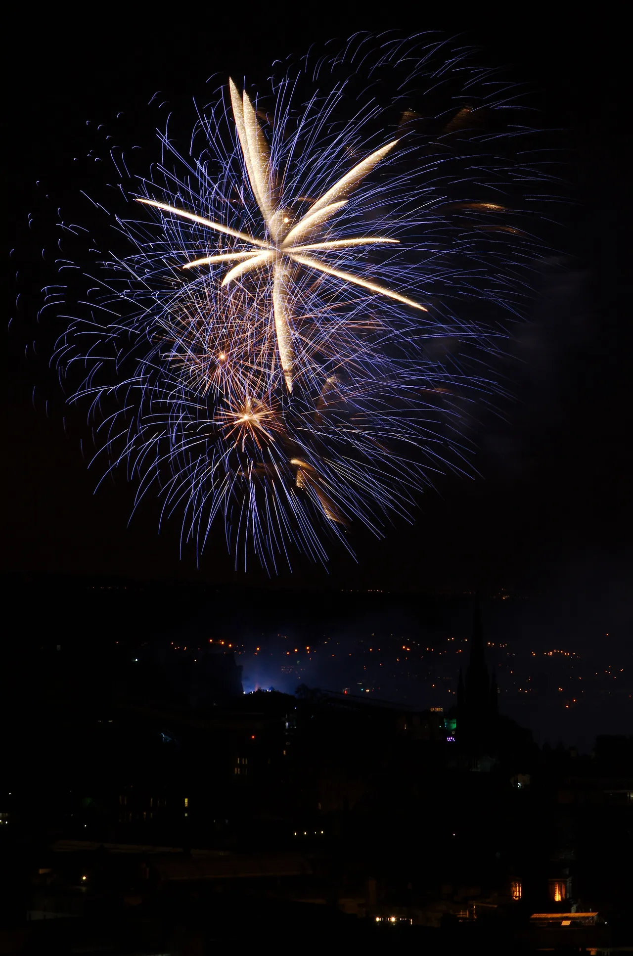 Um fogo de artifício branco e azul sobre Edimburgo na noite de 31 de agosto de 2014, com o castelo e uma torre de igreja no escuro.